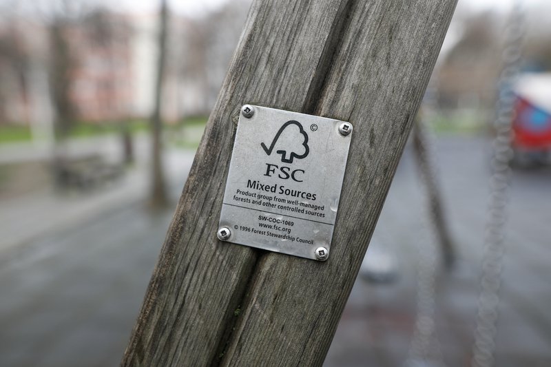 Un primo piano di una struttura in legno con una placca metallica FSC (Forest Stewardship Council) attaccata, che indica l'uso di materiali provenienti da fonti responsabili, con un parco sfocato sullo sfondo.