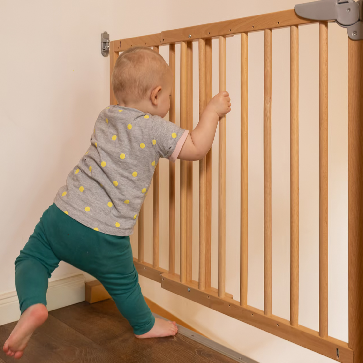 Un bambino si appoggia a un cancelletto di sicurezza in legno fissato al muro, sollevandosi in una stanza con pavimento in parquet.