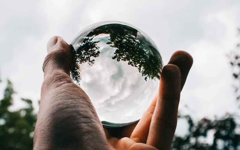Mano che tiene una sfera di vetro riflettente che mostra una vista capovolta degli alberi e di un cielo nuvoloso.